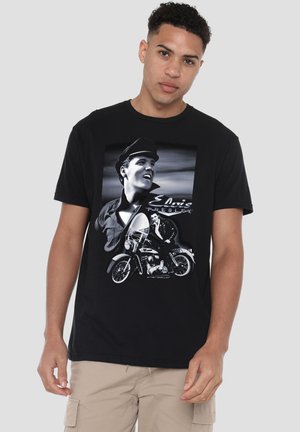 Young man wearing black t-shirt with black-and-white images of Elvis Presley, including a portrait and him sitting on a motorcycle.
