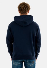 Felpa con cappuccio blu navy, in cotone, vestibilità comoda, polsini e orlo a costine, grande cappuccio sul retro, indossata con jeans azzurri chiari.