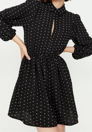 Kobieta ubrana w czarną sukienkę do kolan w drobne białe kropki, z długimi bufkami, zapinanym kołnierzykiem i marszczoną talią.