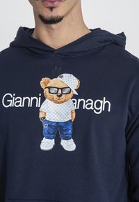 Námořnická modrá mikina s kapucí s karikaturou medvěda ve slunečních brýlích a kšiltovce, bílý text "Gianni" dole, se detailními džíny a botami.