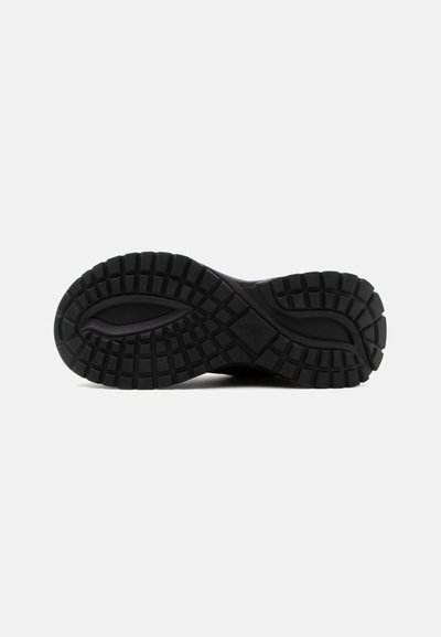 Semelle de chaussure noire avec des motifs de profil profonds multidirectionnels conçus pour l'adhérence et la durabilité, montrée sur un fond clair uni.
