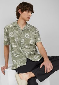 Camicia verde a maniche corte con motivi floreali e geometrici bianchi; tessuto leggero, design con bottoni, orlo dritto. Abbinata a pantaloni neri.