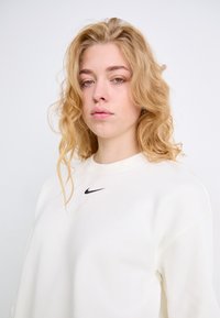 Sudadera color crema con cuello redondo y puños acanalados, con un pequeño logo negro de Nike en el pecho. Textura suave.