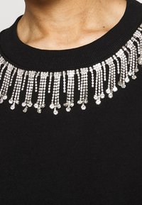 Maglione nero con un frange di strass argento lungo il collo. Il colletto è a costine e liscio, in contrasto con gli accenti scintillanti.