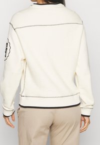 Bluza w kolorze kremowym z prążkowanymi mankietami i dolnym brzegiem, teksturowaną tkaniną oraz kontrastującymi przeszyciami. Na rękawie znajduje się naszywka z logo.