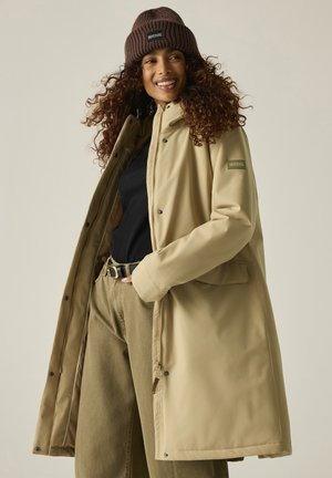 Mujer sonriente con pelo rizado lleva un abrigo largo beige, gorro de punto marrón, camiseta negra y pantalones beige sobre un fondo liso.