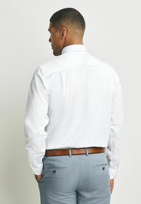 Weißes Langarmshirt von hinten betrachtet, mit klassischem Kragen und geknöpften Manschetten, kombiniert mit hellgrauen Hosen und einem braunen Gürtel.