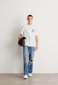 Maglietta bianca con grafica blu sul petto, jeans blu strappati, borsa a tracolla nera e sneakers bianche, posizionata su uno sfondo semplice.