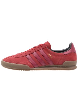 Zapatilla Adidas baja roja con parte superior de ante, tres franjas rojas más oscuras, suela de goma y texto dorado "JEANS" en el lateral.