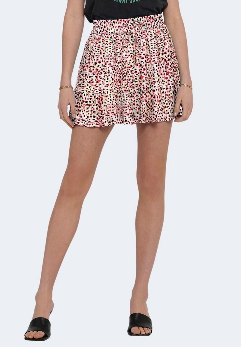 Femme portant une mini-jupe à volants tachetée rose, blanche et noire avec une taille à cordon et des sandales noires à bout ouvert, debout devant un fond clair.