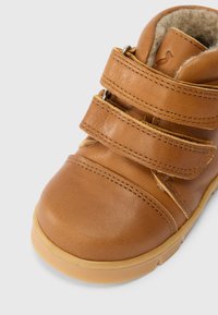 Bota de couro castanho com três tiras de velcro, bico arredondado, sola de borracha bege e forro interior macio para conforto.