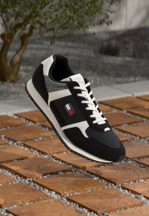 Zwart-witte sneaker met een gestructureerde bovenkant, mesh accenten en witte veters, voorzien van een logo in rood en blauw aan de zijkant, gepresenteerd tegen stenen tegels.