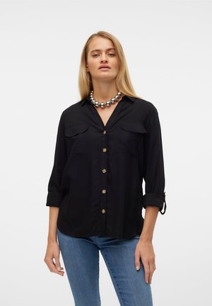 Femme aux longs cheveux blonds portant une chemise noire à boutons avec manches retroussées, un jean bleu et un grand collier de perles argentées sur fond blanc.