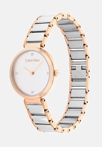Montre Calvin Klein avec un cadran rond blanc, des accents en or rose, un bracelet en acier inoxydable argenté et quatre petits marqueurs en cristal sur le cadran.