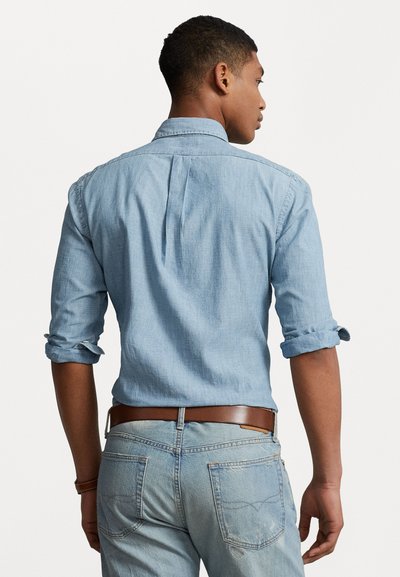 Light blue denim shirt with rolled sleeves, button-down collar, and back pleat, paired with light blue jeans and brown leather belt.