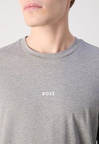 Szürke pamut póló kerek nyakkal, fehér "BOSS" logóval a mellkasán. Az anyag sima, finom textúrával.