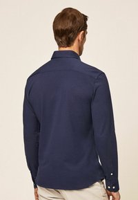 Homme portant une chemise à manches longues bleue foncée à boutons avec un pantalon beige, vu de dos sur un fond uni.