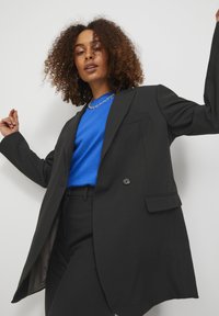 Black blazer made of smooth fabric, featuring a single button, notch lapels, and front pockets, paired with a blue t-shirt underneath.