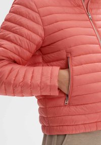 Veste rembourrée en corail avec une poche zippée et une texture côtelée. Elle possède un col montant et une forme ajustée avec de subtils traits de rembourrage horizontaux.