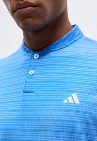 Κοντινή λήψη ενός ατόμου που φορά ένα μπλε αθλητικό πουκάμισο με υφή, μανταρίνι γιακά, δύο κουμπιά και λευκό λογότυπο Adidas στο στήθος.