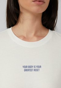 T-shirt en coton blanc avec un col rond. Le devant présente un texte bleu qui dit "VOTRE CORPS EST VOTRE PLUS GRAND BIEN." Texture lisse.