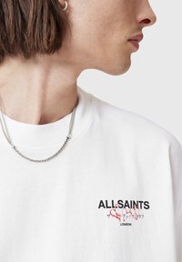 White cotton t-shirt with round neckline, featuring a black and red graphic logo "ALL-SAINTS LONDON" on the left chest. Silver chain necklace visible.