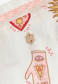 Tessuto decorato con disegni a mano in rosa e oro, con un ciondolo dorato attaccato, il tutto su uno sfondo bianco testurizzato.