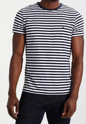 Man draagt een zwart-wit horizontaal gestreept T-shirt met korte mouwen en een donkere broek, staand met ontspannen armen.