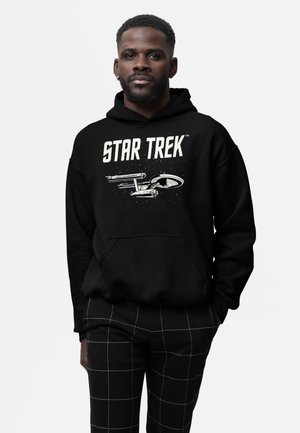 Mężczyzna ubrany w czarną bluzę Star Trek z grafiką statku kosmicznego oraz czarne spodnie z białymi liniami w kratkę, stojący na jednolitym tle.