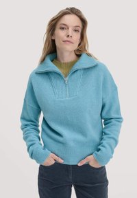 Helles blaues Zip-up Sweatshirt mit geripptem Kragen und Bündchen, ausgestattet mit einem strukturierten Vorderteil. Model trägt dunkle Cordhosen.