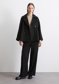 Manteau en laine noir avec des boutons à double boutonnage, grand col et manches larges, associé à un pantalon noir fluide et des chaussures noires.