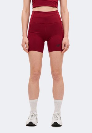 Le mannequin porte un short de sport bordeaux taille haute et un crop top assorti, avec des chaussettes blanches et des baskets, et se tient devant un arrière-plan uni.