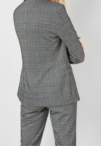 Personne portant un blazer à carreaux gris et un pantalon assorti, bras croisés, vue de dos sur un fond neutre.