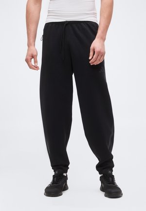 Mężczyzna ubrany w czarne spodnie dresowe Adidas z elastycznymi ściągaczami, czarne sportowe buty i biały top bez rękawów, stojący na jednolitym tle.
