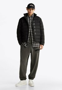 Czarna kurtka puchowa z logo, koszula w kratę, szare dresy z napisem i białe trampki. Luźny, warstwowy strój.