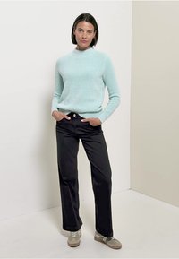 Hellblauer Strickpullover mit hohem Kragen, kombiniert mit schwarzen Hosen mit weitem Bein. Das Modell trägt hellgraue Sneakers. Einfache, legere Kleidung.