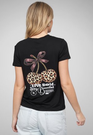 Kobieta o blond włosach nosi czarną koszulkę z nadrukiem czereśni w panterkę związanych fioletową kokardą oraz napisem „Live bold stay beautiful” na plecach.