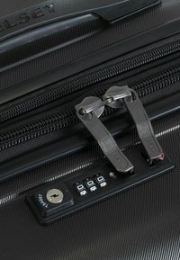 Valigia nera con guscio rigido, cerniera con doppie maniglie metalliche argentate, combinazione di lucchetto e chiave in plastica. Superficie testurizzata con scanalature parallele.