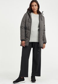 Dunjakke i mørkegrå med hette og quiltet design, kombinert med en lys grå genser, svarte vide bukser og svarte joggesko.