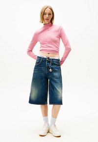 Rosa Langarmshirt, blaue Denim-Culottes, weiße Sneakers und hellgraue Socken. Hervorzuhebende Details: Logo auf dem Shirt, Schlüsselanhänger an der Jeans.