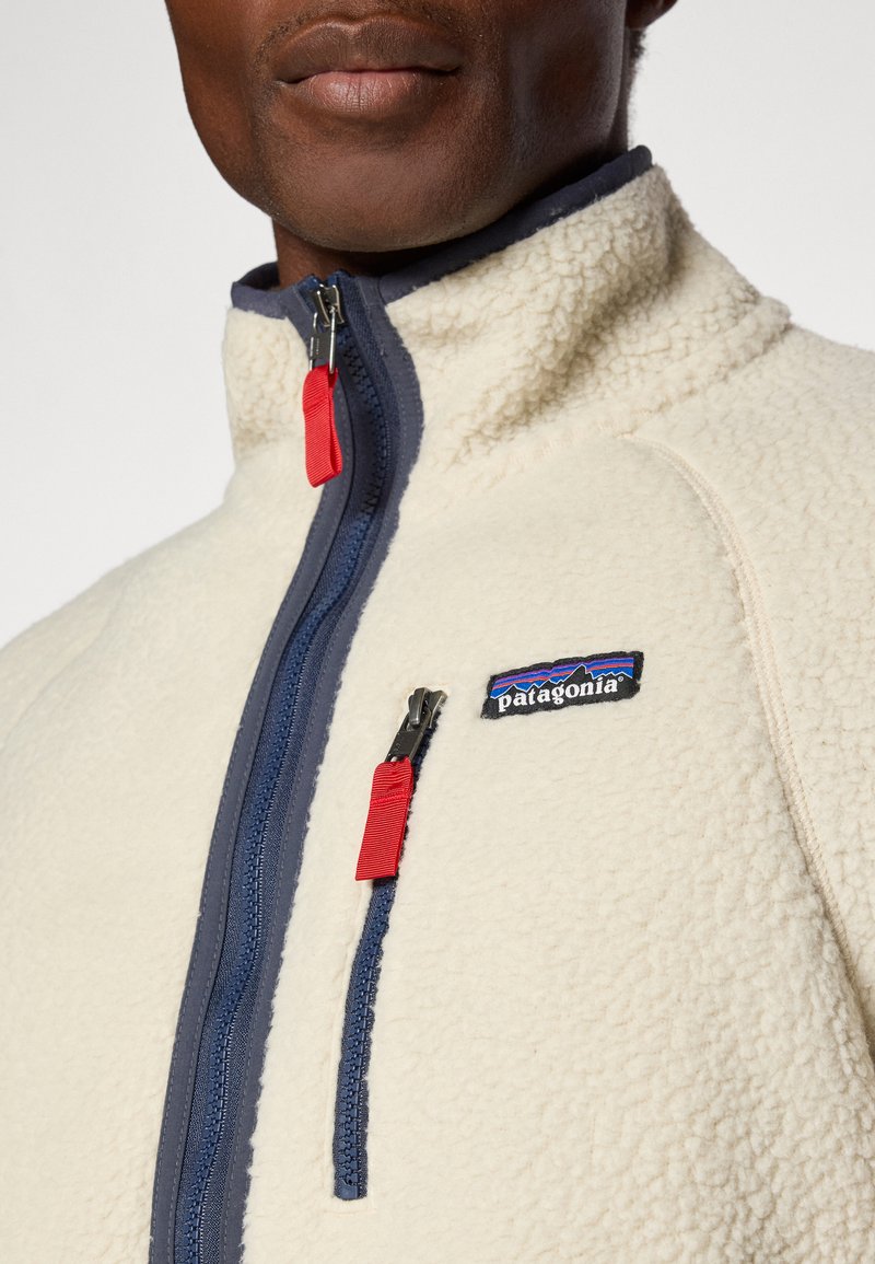 Giacca in pile color crema con colletto alto, dettagli blu navy e zip rosse. Presenta una tasca sul petto con il logo Patagonia.