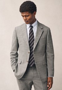 Completo grigio a quadri con vestibilità su misura, caratterizzato da due bottoni, rever a lancia e pantaloni coordinati. Sotto, una camicia azzurro chiaro con piccoli puntini e una cravatta a righe.