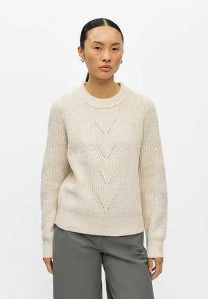 Femme aux cheveux noirs raides portant des boucles d’oreilles créoles dorées, un pull en maille texturé crème avec motif chevron, et un pantalon gris sur fond blanc.