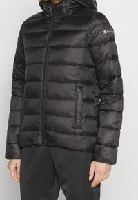 Svart vadderad jacka med quiltad design, hel dragkedja framtill och huva. Tillverkad av glansigt material med sidofickor och logotypdetalj.