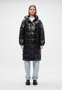 Melnā, spīdīgā pūkainā jaka ar kapuci, ar rāvējslēdzēju un ribotu apakšmalu. Apvienota ar gaiši zilām džinsām un melniem masīviem zābakiem.