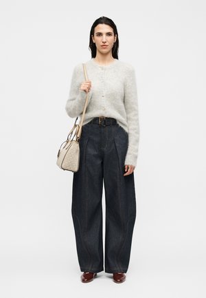 Vrouw draagt een lichtgrijze, zachte cardigan, donkere wijde broek met een riem, bruine schoenen en een beige schoudertas tegen een effen achtergrond.