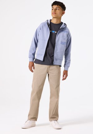 Felpa con zip di colore azzurro chiaro, con tasche frontali e testo stampato, indossata sopra una maglietta scura, abbinata a pantaloni beige e sneakers bianche.