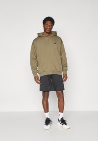 Olive green zip-up hooded sweatshirt with a large front pocket, paired with black shorts and black and white athletic shoes.