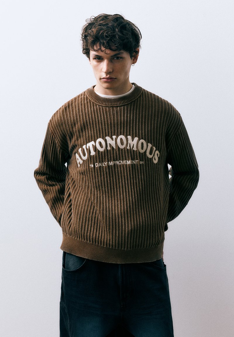 Pull marron côtelé avec les inscriptions "AUTONOMOUS" et "DAILY IMPROVEMENT" en crème sur la poitrine. Encolure ronde et coupe décontractée.