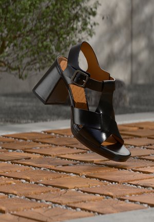 Sandale à talon en cuir noir avec bride à la cheville et boucle reposant sur un sol en carrelage en bois à l'extérieur.